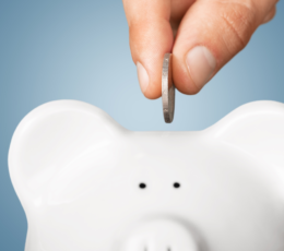Close-up of a hand inserting a coin into a white piggy bank against a blue background to symbolize saving money